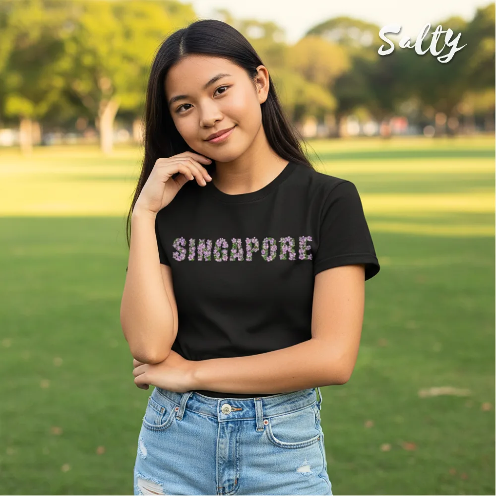 Black t-shirt with bold floral SINGAPORE print across chest, styled with light blue denim shorts in outdoor park setting. Wet Tee Shirt® x Salty collaboration highlighting location-inspired streetwear fashion for casual urban style.