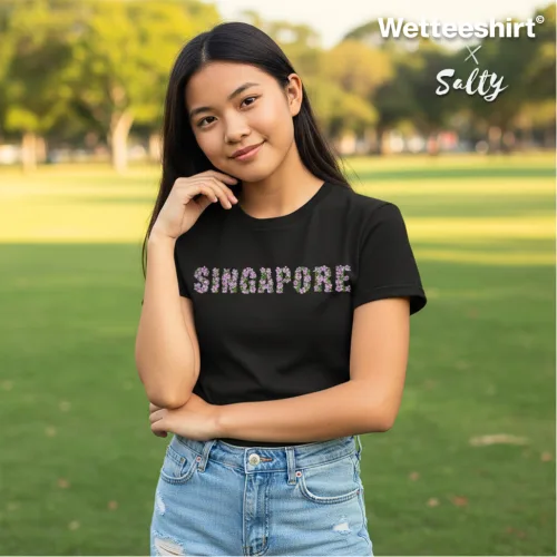 Black t-shirt with bold floral SINGAPORE print across chest, styled with light blue denim shorts in outdoor park setting. Wet Tee Shirt® x Salty collaboration highlighting location-inspired streetwear fashion for casual urban style.