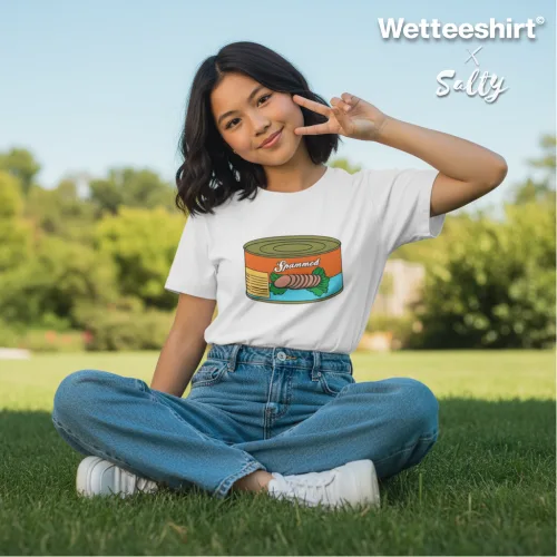 Casual white graphic t-shirt with colorful canned food design, worn by smiling person sitting cross-legged on grassy field. Playful streetwear fashion lifestyle shot with peace sign gesture, trees in background. Wet Tee Shirt® brand promotional image showcasing quirky, unique apparel for trendy youth culture.
