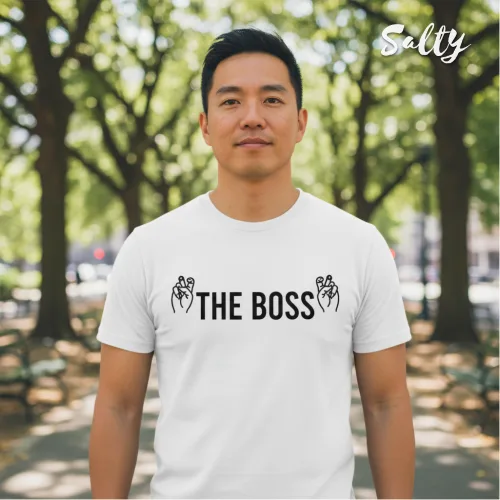 Singapore streetwear — man outdoors on tree‑lined park pathway wearing Wetteeshirt® x Salty white graphic t-shirt with bold black ‘THE BOSS’ text flanked by hand gesture illustrations, casual lifestyle fashion shot with greenery and benches in background.