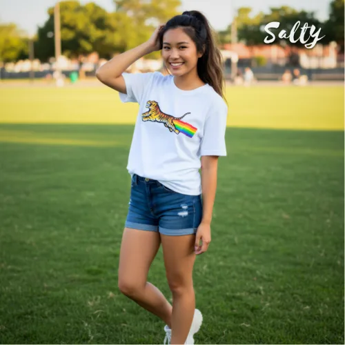White graphic t-shirt featuring bold tiger design breathing rainbow, styled with denim shorts and white sneakers. Outdoor streetwear fashion photo on grassy sports field with casual vibe. Wet Tee Shirt x Selly collaboration showcasing unique, playful apparel for trendy youth culture.
