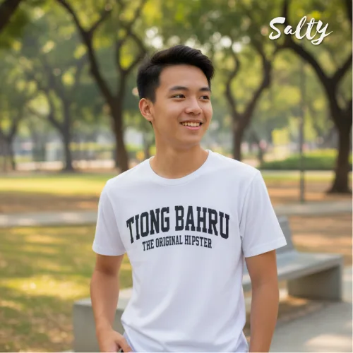 Singapore streetwear — young person outdoors in park with trees and benches wearing Wetteeshirt® × Salty white graphic t-shirt with bold black ‘TIONG BAHRU THE ORIGINAL HIPSTER’ text, casual lifestyle fashion shot with greenery and urban backdrop.