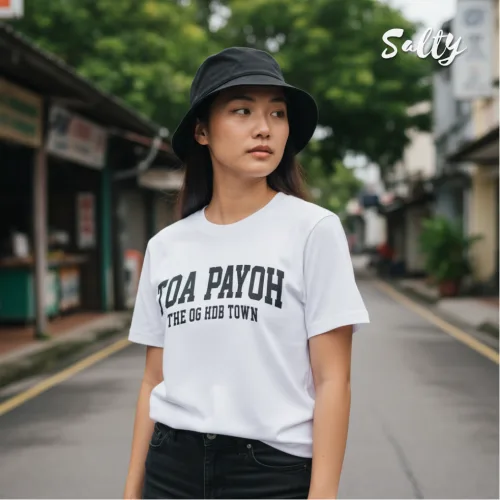 Streetwear photo of a person in a black bucket hat and white graphic tee reading ‘TOA PAYOH THE OG HOOD TOWN,’ shot on a quiet Singapore street with greenery and low‑rise buildings, part of the Wet Tee Shirt® x Salty local culture collection.