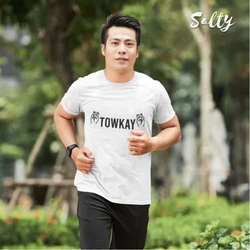 Singapore streetwear — man jogging outdoors in park wearing Wetteeshirt® x Salty white graphic t-shirt with bold black ‘TOWKAY’ text flanked by hand gesture illustrations, styled with black athletic pants and wristwatch, casual lifestyle fashion shot with greenery and blurred pathway background.