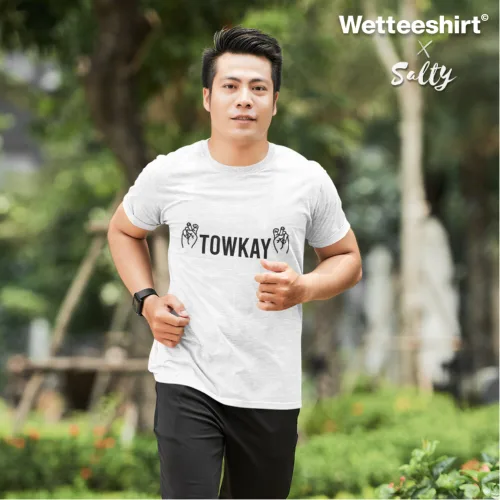 Singapore streetwear — man jogging outdoors in park wearing Wetteeshirt® x Salty white graphic t-shirt with bold black ‘TOWKAY’ text flanked by hand gesture illustrations, styled with black athletic pants and wristwatch, casual lifestyle fashion shot with greenery and blurred pathway background.