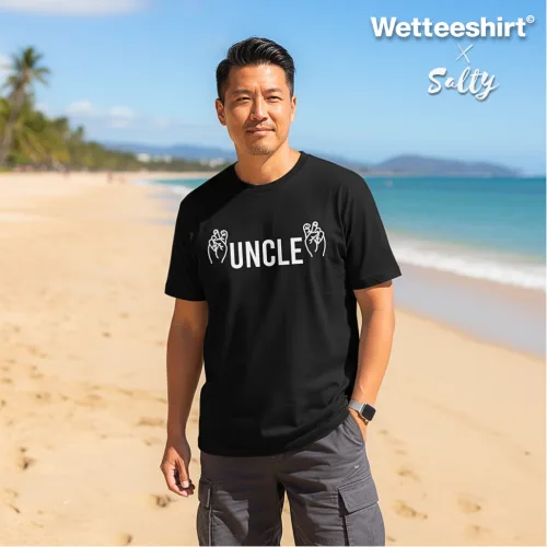 Black UNCLE graphic t-shirt with bold white lettering and rock hand gesture icons, styled with gray cargo shorts on tropical sandy beach with palm trees and ocean backdrop. Wet Tee Shirt® x Salty collaboration showcasing playful lifestyle streetwear fashion for casual island vibes.