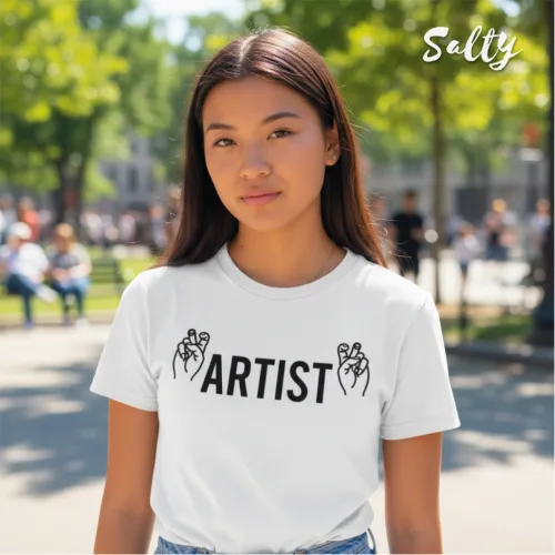 Singapore streetwear — person outdoors in park wearing Wetteeshirt® x Salty white graphic t-shirt with bold black ‘ARTIST’ text flanked by expressive hand gesture illustrations, casual lifestyle fashion shot with trees, grass, and people in background.
