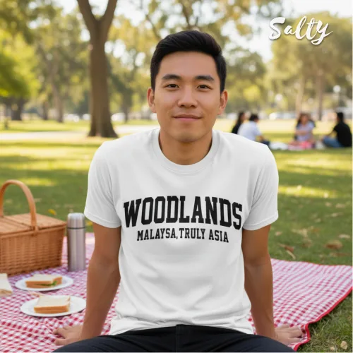 Singapore streetwear — person outdoors on red checkered picnic blanket wearing Wetteeshirt x Selly white graphic t-shirt with bold black text ‘WOODLANDS MALAYSIA, TRULY ASIA,’ casual lifestyle fashion shot with wicker picnic basket, sandwiches, cake, and park trees in background.