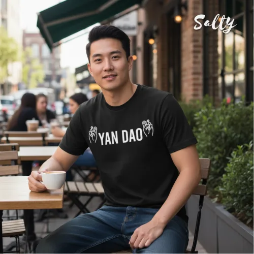Casual urban photo of a man at an outdoor café holding a cup, wearing a black graphic tee with bold white text ‘YAN DAO’ and hand illustrations, surrounded by tables, sidewalk, and buildings, part of the Wet Tee Shirt® x Salty Singapore streetwear collection.