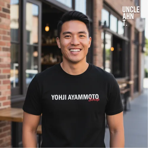 Streetwear photo of a smiling man in a black graphic tee with bold white and red text ‘YOHJI AYAMMOTO,’ standing outside a brick café with large windows, part of Wet Tee Shirt® x Salty Singapore’s Uncle Ahn T collection.