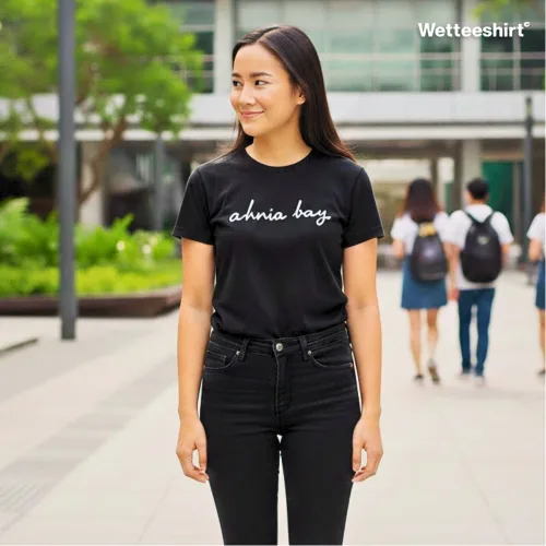 Person in black Wetteeshirt® Ah Nia Bay tee with cursive logo, casual unisex streetwear in city setting.