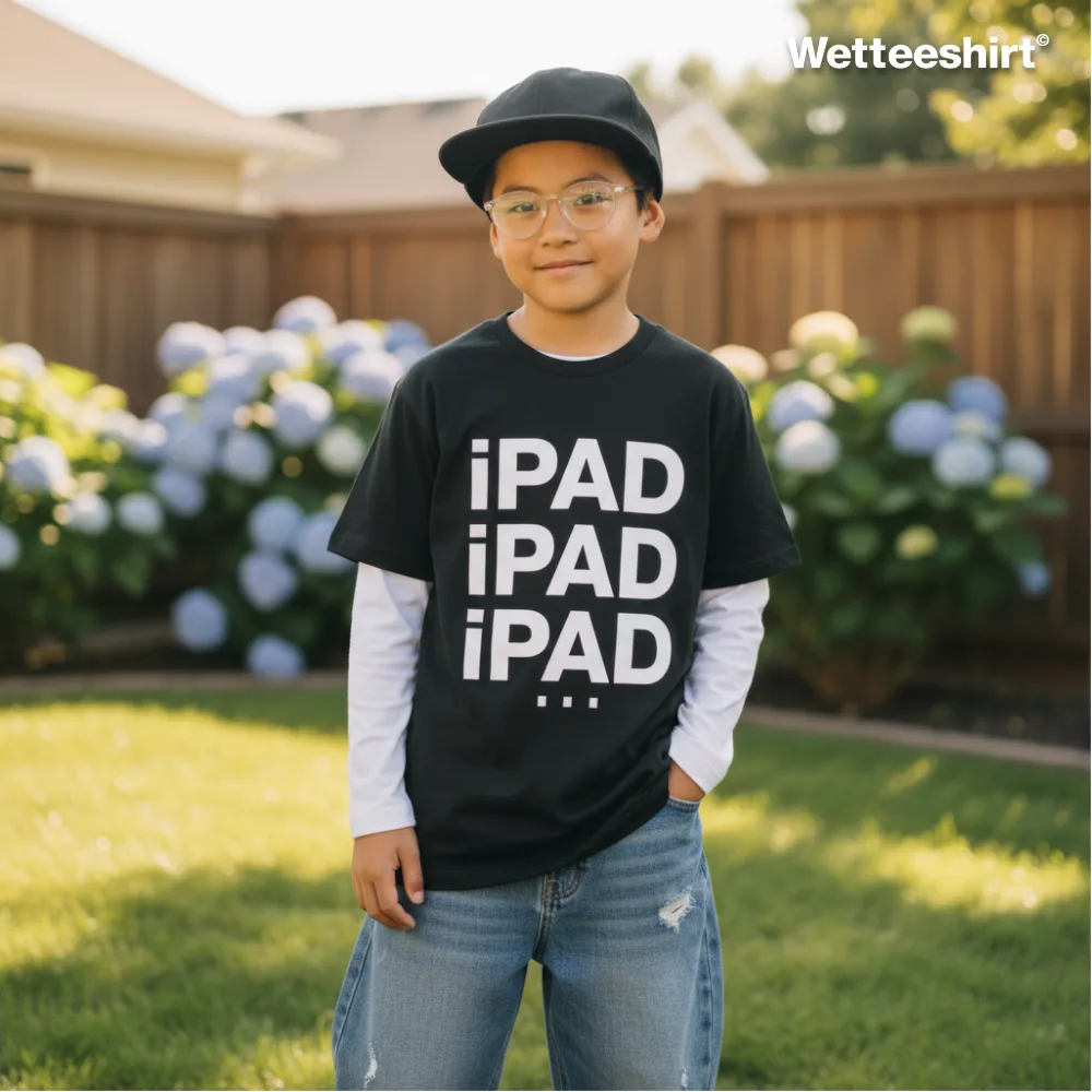 Black casual T-shirt with bold vertical white ‘¡PAD ¡PAD ¡PAD…’ slogan, styled over long-sleeved white shirt with blue jeans, black cap, and glasses, worn outdoors in backyard with wooden fence and blooming hydrangeas — witty streetwear parody inspired by Apple iPad branding.